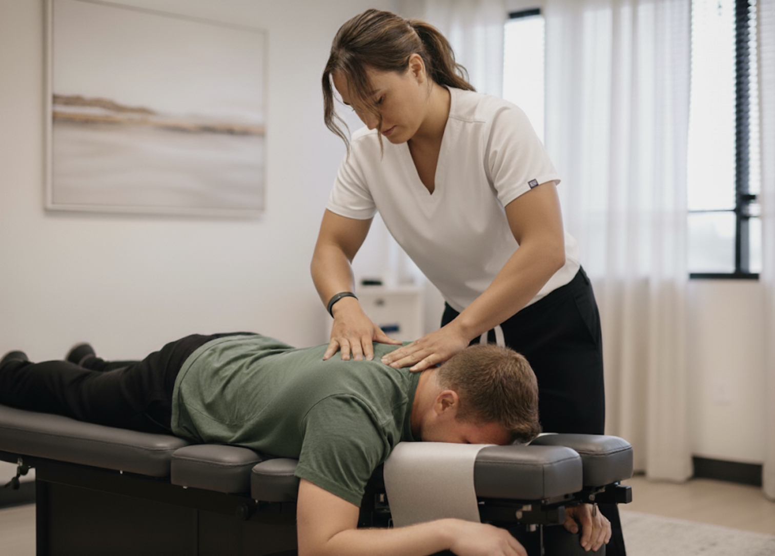 A chiropractor in a white uniform is performing a back adjustment on a man lying face down on a massage table. The man is wearing a green shirt and appears relaxed in a well-lit room with white curtains and a landscape painting on the wall.