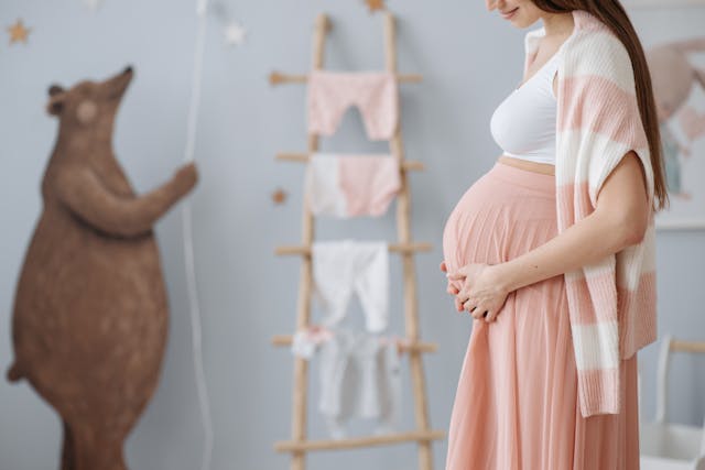 Image of a pregnant woman in a pink skirt, pink and white sweater over her shoulders and white shirt standing in a nursey holding her pregnant belly. 