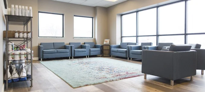 Image of the office waiting room. The room is well lit with a lot of windows, blue chairs and a rug in the middle is a light green. 