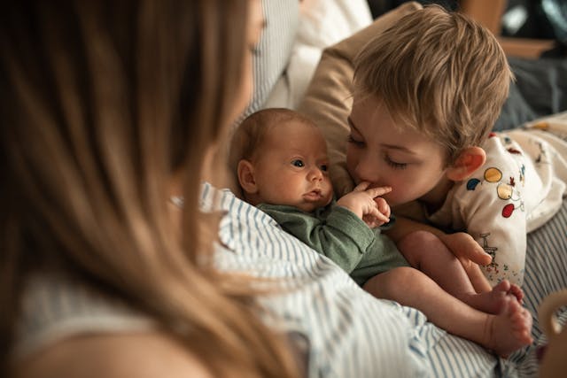 Image of a mom looking down at her baby and young child in her lap. The young boy is kissing the baby's hands. 