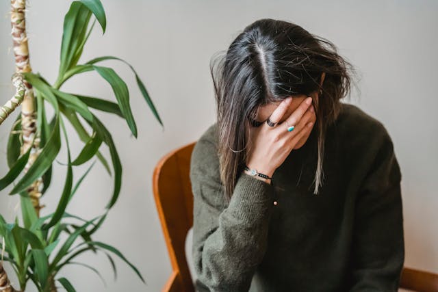 Image of a woman with her head down and covered by her hand. The background is a grey wall. There is a plant in the background and the woman is sitting on a wooden chair. 