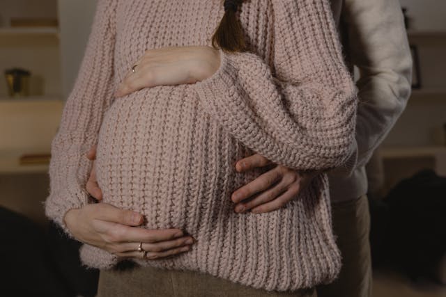 A close-up of a pregnant belly, with a man's arms wrapped around the woman as they both gently hold the belly.