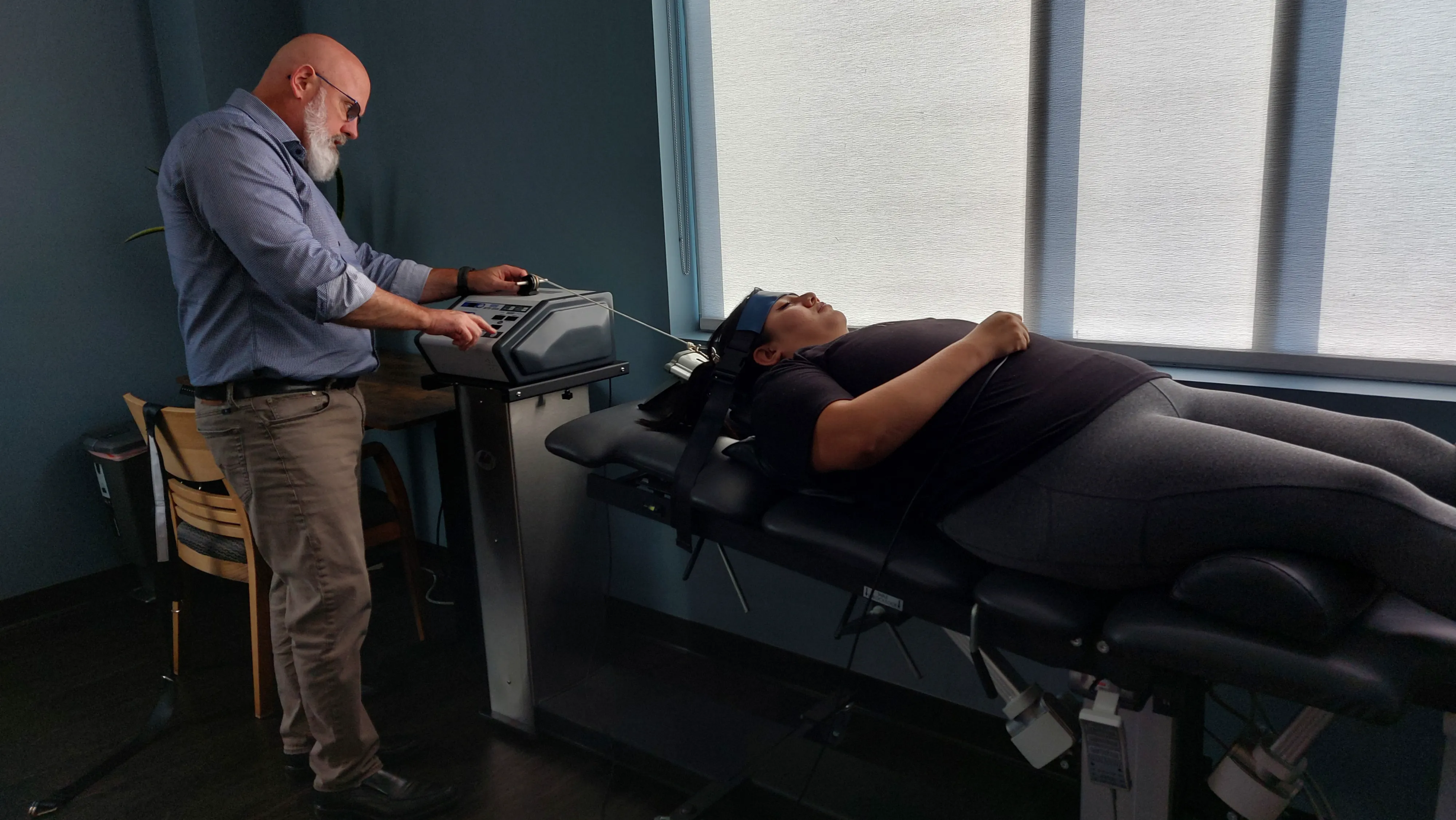 A woman in gray pajamas sits on the edge of a bed, pressing her hands against her lower back in discomfort. The room is softly lit, with a glass of water on the bedside table and a neatly arranged bed in the background.