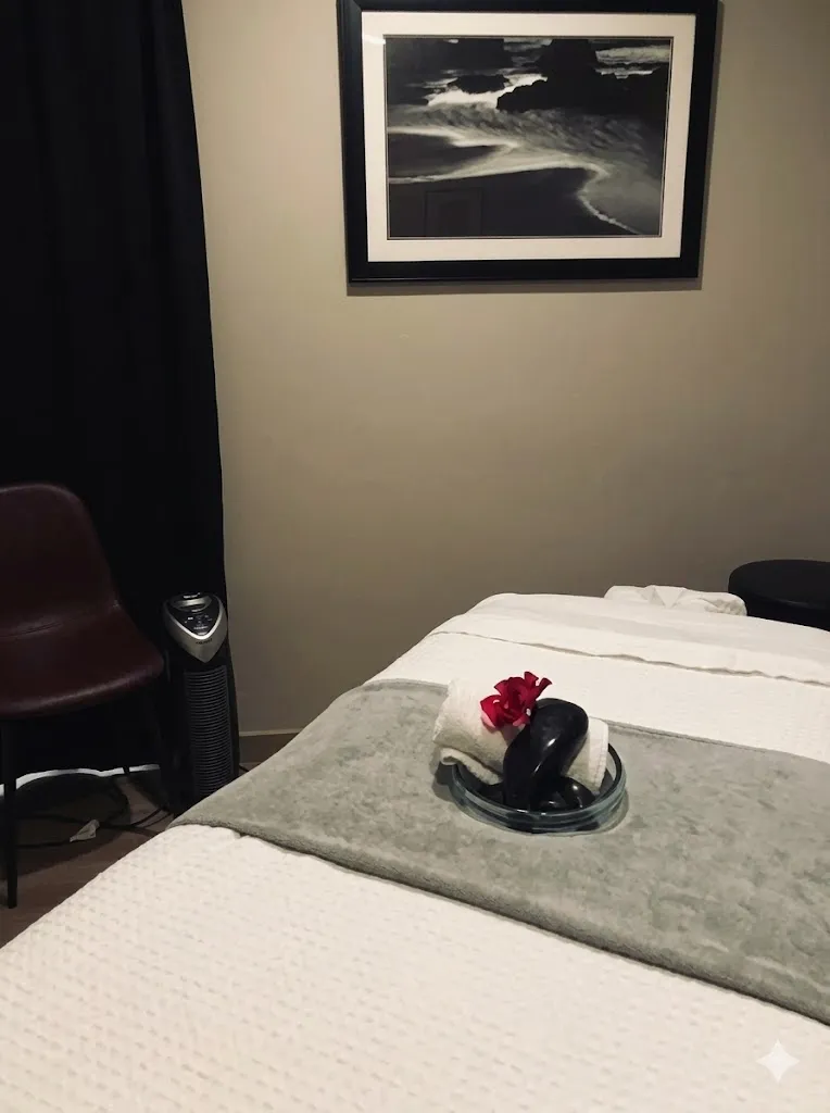 The serene massage therapy treatment room at The Back and Neck Pain Center of Monterey, CA. The image features a prepared massage table with quilted bedding, a central bowl containing hot stones, a folded towel, and a vibrant red flower, alongside framed coastal art on a tan wall, an air purifier, and a dark leather chair. We provide a calming environment for holistic recovery.
